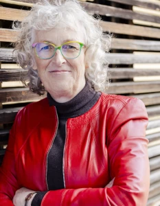 Martina Kreutz mit roter Lederjacke und braunem Rolli an eine Holzfassade gelehnt