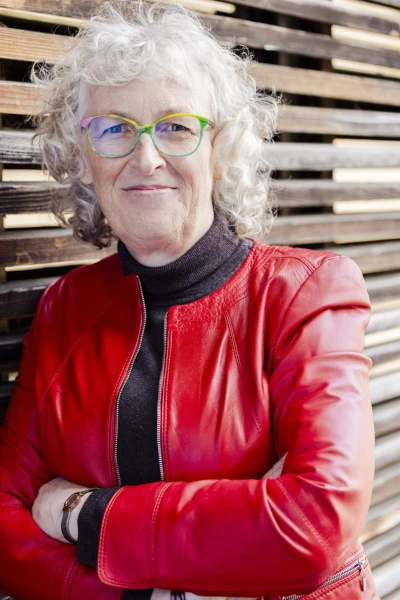 Martina Kreutz mit roter Lederjacke und braunem Rolli an eine Holzfassade gelehnt