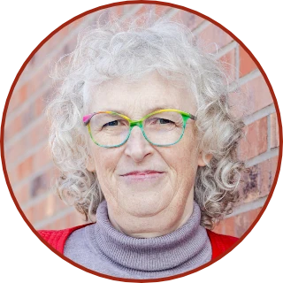 Martina Kreutz mit roter Lederjacke und grauem Rolli an eine Klinkerfassade gelehnt